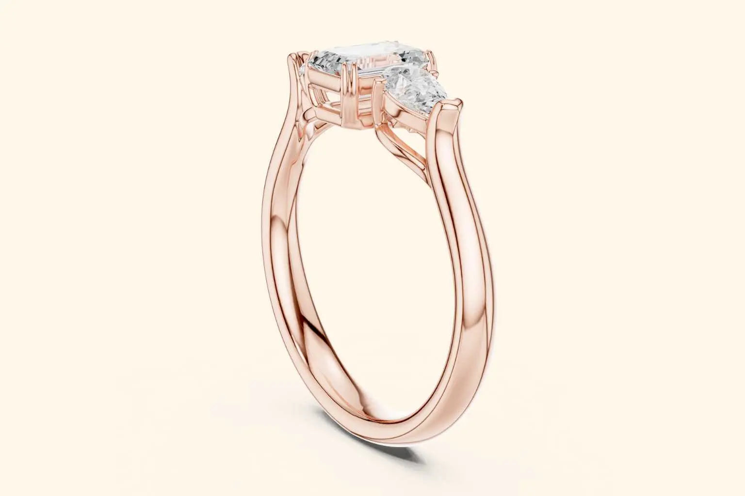 A side-profile view of a three-stone ring in polished rose gold. It clearly shows the slightly raised setting of the central emerald-cut diamond and one pear-shaped side diamond, with the narrow band curving underneath.