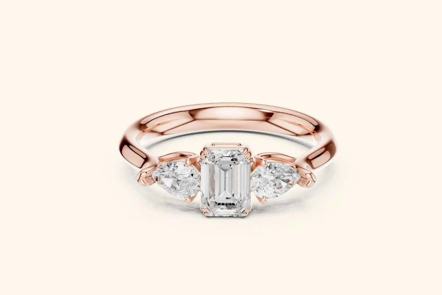 A front view of a statement three-stone ring in brilliant rose gold. The design showcases a horizontal emerald-cut diamond in the center, perfectly balanced by two sparkling pear-shaped diamonds on the shoulders.