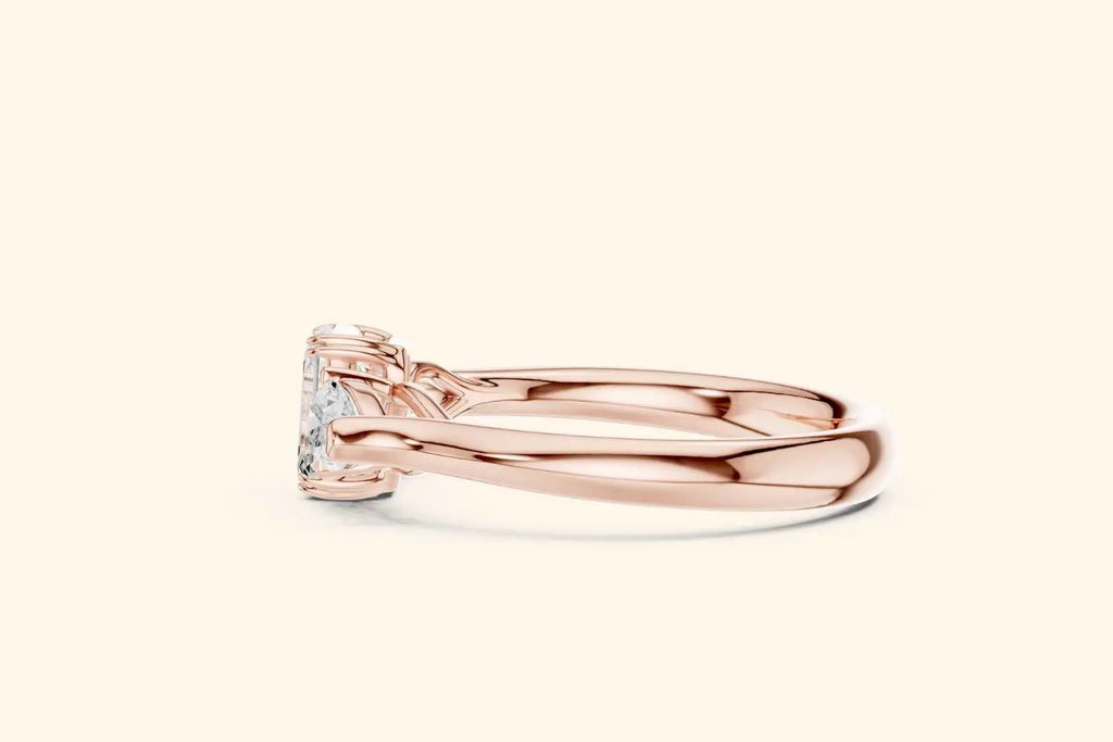 A close-up horizontal side view of a wide-band three-stone ring in polished rose gold. The view emphasizes the low-profile setting where the emerald and pear-cut diamonds sit close to the finger.